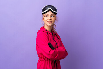 Skier teenager girl with snowboarding glasses over isolated purple background with arms crossed and happy