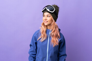Skier teenager girl with snowboarding glasses over isolated purple background looking to the side and smiling