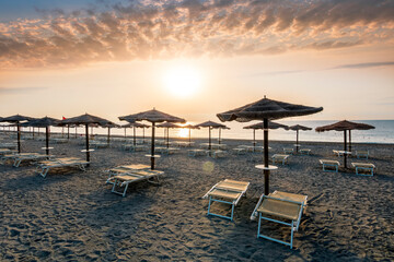 beautiful landscape of empty sunrise or sunset beach with chaise loungues and nice umbrellas with blue sea, sun glow and amazing cloudy sky on thr background