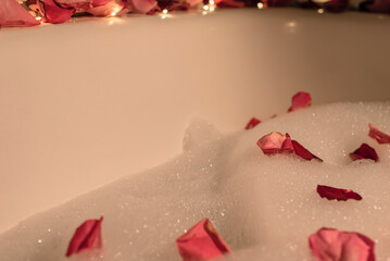 Bathtub with foam, pink rose petals and lights. Enjoying life, luxury, love concept. Valentine or other holiday greeting card with copy space