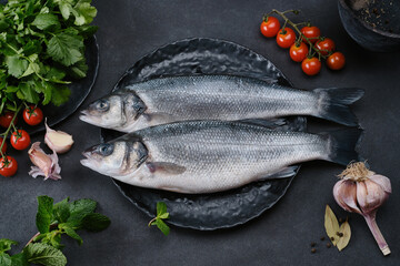 Fresh raw fish Seabass on a black plate with spices. Two fresh Sea Bass fishes with garlic, mint,...