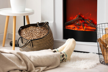 Bag with wood pellets and woman warming near fireplace at home