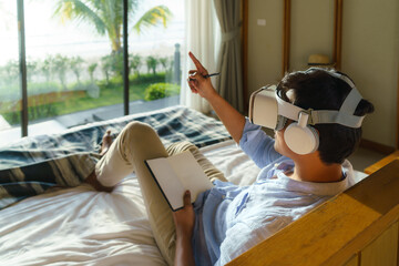 Young Asian man lying on bed and using virtual reality glasses for business meeting during his travel summer vacation holiday.