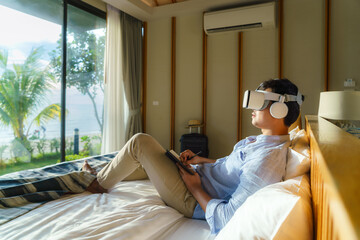 Young Asian man lying on bed and using virtual reality glasses for business meeting during his...