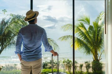Asian man with a suitcase stand near the window of his room and looking at the beautiful sea view...