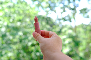 close-up female hand holding a tube with new pink, peach lipstick on natural background of summer foliage, wants to do makeup, concept of natural cosmetics, power of nature for female beauty