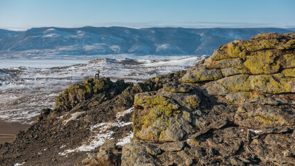 Yellow lichens grow on cracked granite rocks. At the top of the cliff there is a man with his hand raised up. In the distance - mountains against a blue sky, a frozen lake. Baikal
