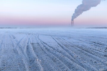 Winter minimalistic landscape with snowmobile prints in the snow and smoking chimneys on the horizon.