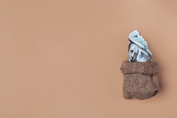 Roll of one hundred dollar bills in a burlap sack on a beige background. Copy space
