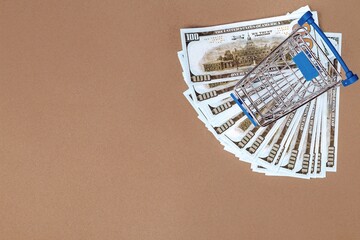 Shopping cart on a fan of one hundred dollar bills on a beige background. Copy space