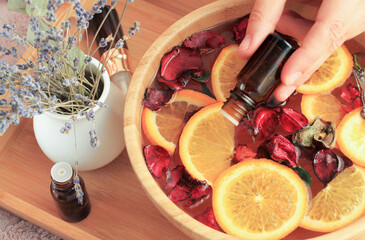 Herbal spa with essential oils. Woman hand pouring drops of aroma extract oil from bottle to bowl with mix flower petals and citrus orange slices. Closeup relaxing lavender botanical spa, calming warm