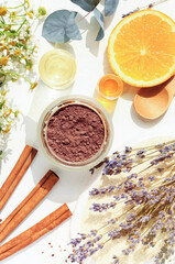 Making homemade skincare treatment facial mask cosmetic clay chocolate powder,  natural ingredients cinnamon orange essential oil, holistic herbs lavender fresh chamomile twigs, top view sunny table