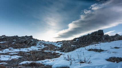 A hill against a background of blue sky and clouds. Snow on the ground. Dry grass among rocks and in snowdrifts. Siberia