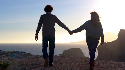 Fototapeta premium man and woman walk on a cliff by the sea at sunset in total freedom of nature - they spread their arms holding hands, kiss and hug during the journey in Murcia Spain La Manga