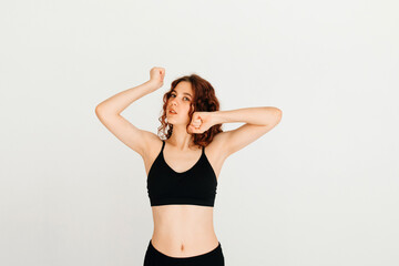 Fototapeta na wymiar Young redhead yoga instructor stretching her wrists, wearing black top, standing at white background, looking at the camera. Morning exercise