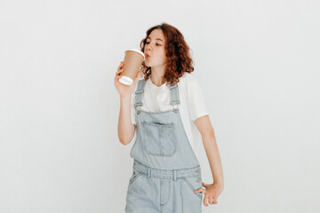 Cheerful redhead girl, wearing denim dungarees and white t-shirt, drinking hot beverage from a coffee cup, glad to finish studying over white background