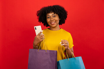 Fototapeta na wymiar Smiling African American woman in casual posing with piles of paper shopping bags and holding blank card isolated over red background.