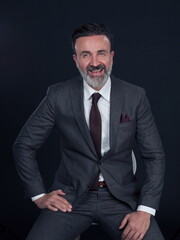 Portrait of adult businessman wearing trendy suit and sitting in modern studio on stylish chair against the black background