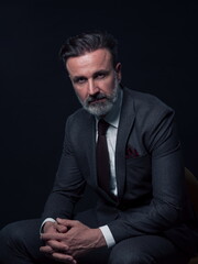Obraz premium Portrait of adult businessman wearing trendy suit and sitting in modern studio on stylish chair against the black background