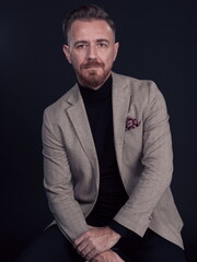 Portrait of adult businessman wearing trendy suit and sitting in modern studio on stylish chair against the black background