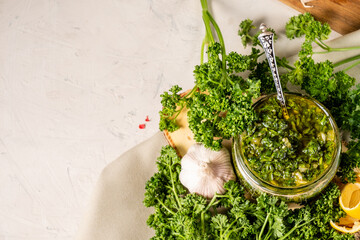 Homemade green chimichurri sauce in a glass jar on a background of parsley, garlic, white table, free space.