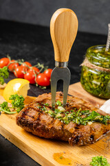 Grilled beef steak on a wooden board, on a background of cherry tomatoes, chemichurri sauce. A chopping fork is in the meat.