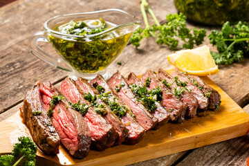Appetizing beef steak cooked and sliced on a wooden table with chimichurri sauce. - obrazy, fototapety, plakaty