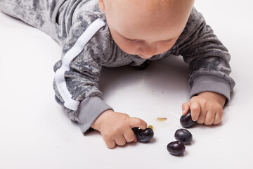 Charming little baby boy in grey clothes eating first food black grape on white background