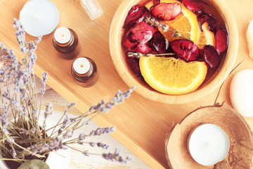 Top view wooden bowl with mix of rose petals and orange citrus slices, essential oils dropper bottles, lavender twigs. Home aromatherapy spa beauty treatment. Soft pink light pastel color.
