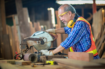 Asian elderly carpenter craftsman in carpentry shop use circular saws to cut wood board to make furniture. And wear safety equipment at work such as gloves, noise-canceling, headphones.