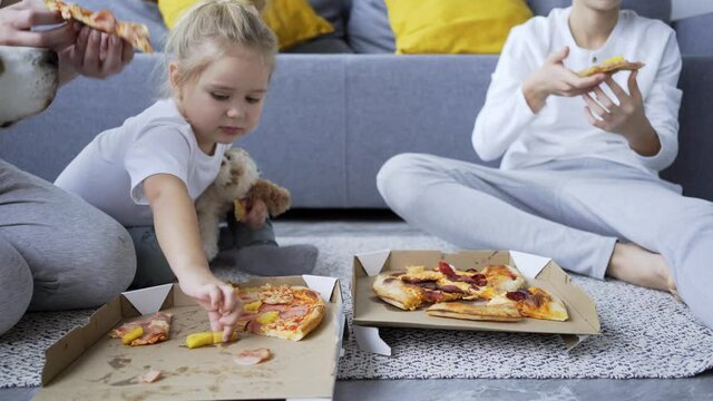 Close-up young woman with little daughter and teenage blue haired son eats tasty pizza near couch sitting on floor in living room at home, detail view box and hands takes pices of pizza