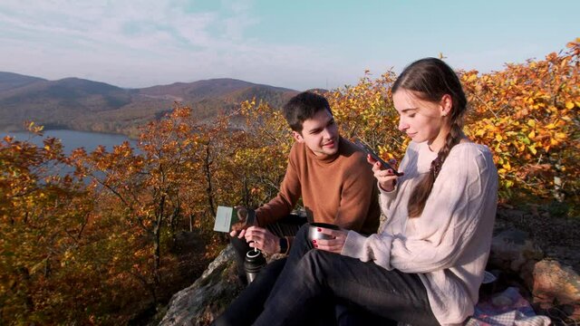 A couple is sitting on the big rock on the mountain peak, drinking hot tea and making video of the autumn landscape, blue river far away under the bright sun, and a cup in girl's hand