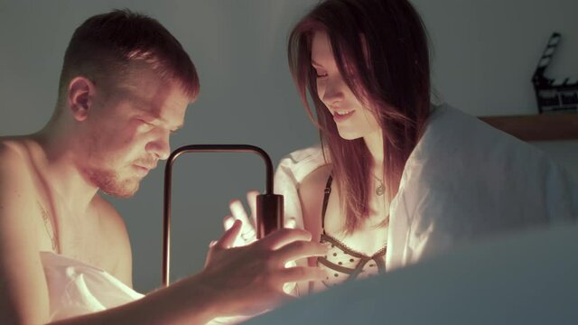 A man and a woman are playing with a lamp in bed. They move their hands around the light. Spend leisure time in the bedroom. Shadows move over the side.