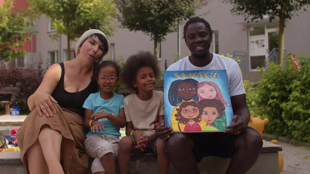 Black man and caucasian woman with biracial children posing looking at camera, happy smiling, waving hand. The father is holding a picture of a family portrait. Forming and performing racial identity.