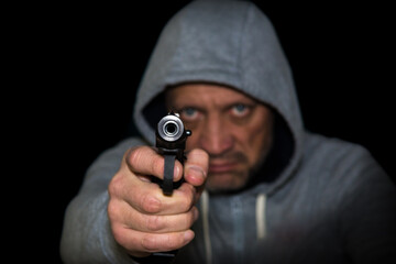 Hooded robber armed with a pistol on a black background. Armed robbery, focus in the foreground. The man is aiming with a pistol.