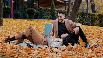 Date outdoors young bearded arab guy sincerely talking with beloved woman man nods approvingly beautiful girl lying on plaid in yellow leaves reading book happy couple smiling together in autumn park