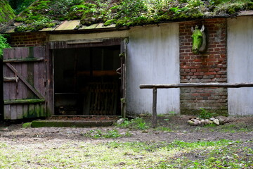 Fototapeta na wymiar A view of old, abandoned stables or barn with the figure of a horse on them, wooden and brick elements, as well as wooden doors opened ajar seen in the middle of a dense forest in Poland