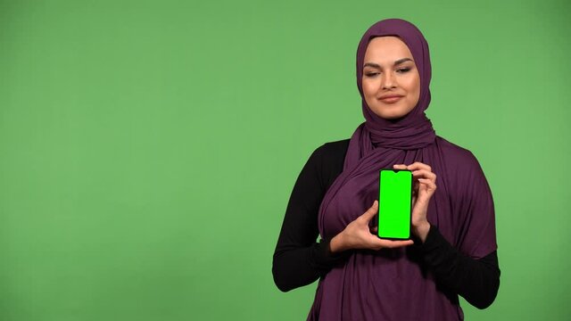 A young beautiful Muslim woman shows a smartphone with green screen to the camera with a smile - green screen background