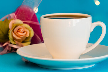 Scena con una tazzina bianca di caffè sul piattino, con dietro un cuore rosa con un fiore sopra, su sfondo azzurro con lucine bianche. Colazione. Mattina. Pausa caffè. Still life.