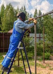 Man welder in a welding mask, construction uniform and protective gloves cooks metal on a street construction site. Construction of a pavilion, pergola near a country house on a summer day.
