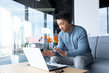 Happy and smiling Asian man in a modern office uses a headset for a video call, a businessman smiles at the interlocutor, and looks at the laptop screen