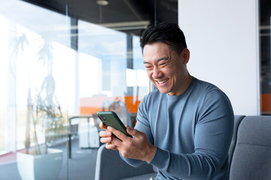 Successful and happy asian businessman man celebrating victory sitting and working in modern office at desk, celebrating victory looking at camera and joyfully shouting holding mobile phone