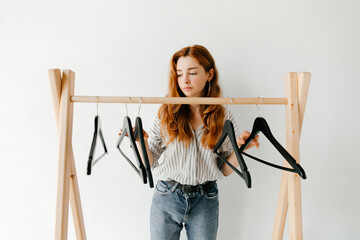 Beautiful disappointed young woman with empty clothes hangers on wooden clothes rack, minimalistic consumption of goods concept.