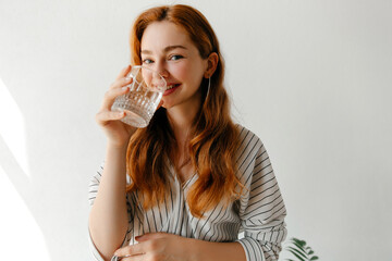 Portrait of smiling redhead woman maintaining her water balance, drinking a glass of pure water,...