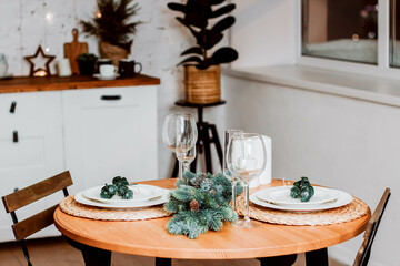Round table decorated for Christmas and New Year. Christmas tree twigs on the table. Plates and wine glasses on the table. Kitchen set and flowers on the background. Christmas content.