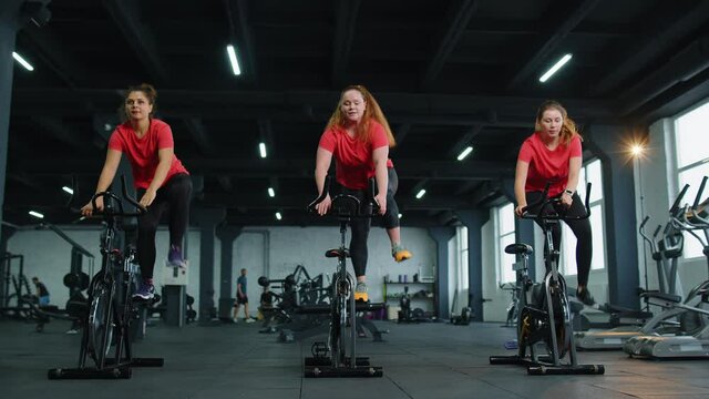 Class group of young women exercising on stationary cycle machine bike in gym. Modern sport activity, workout, healthy lifestyle. Athletic girls spin bike, making tricks indoors. Slow motion