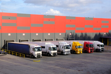 Row of several colorful trucks, side by side, in front of huge logistic center, warehouse