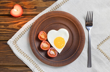 top view of scrambled eggs in the shape of a heart, a symbol of love, fried egg on a clay plate with tomatoes, on a wooden background. Template for a healthy breakfast, close-up.