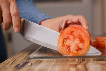 person cutting red tomato with a knife on wooden table in his kitchen in the comfort of his home