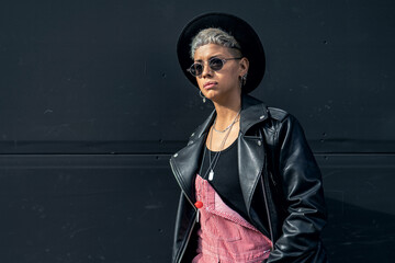 Fashionable modern young woman wearing sunglasses, black hat and jacket, posing on the black city...